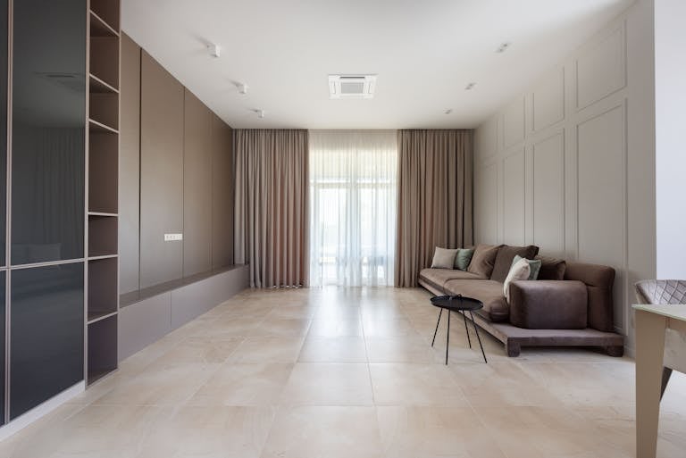 Interior of bright modern flat with sofa with pillows near round table next to cabinet with shelves and curtains on windows